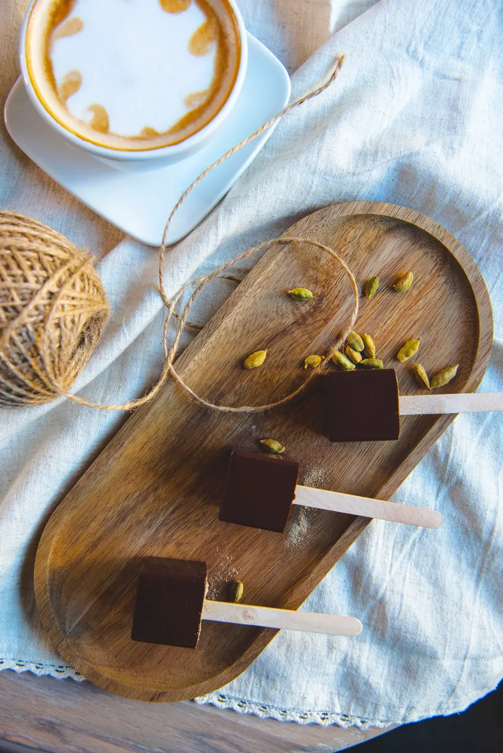 Ciocolată caldă neagră cu cardamon Choco Toni pe băț, pe un platou de lemn lângă un cappuccino