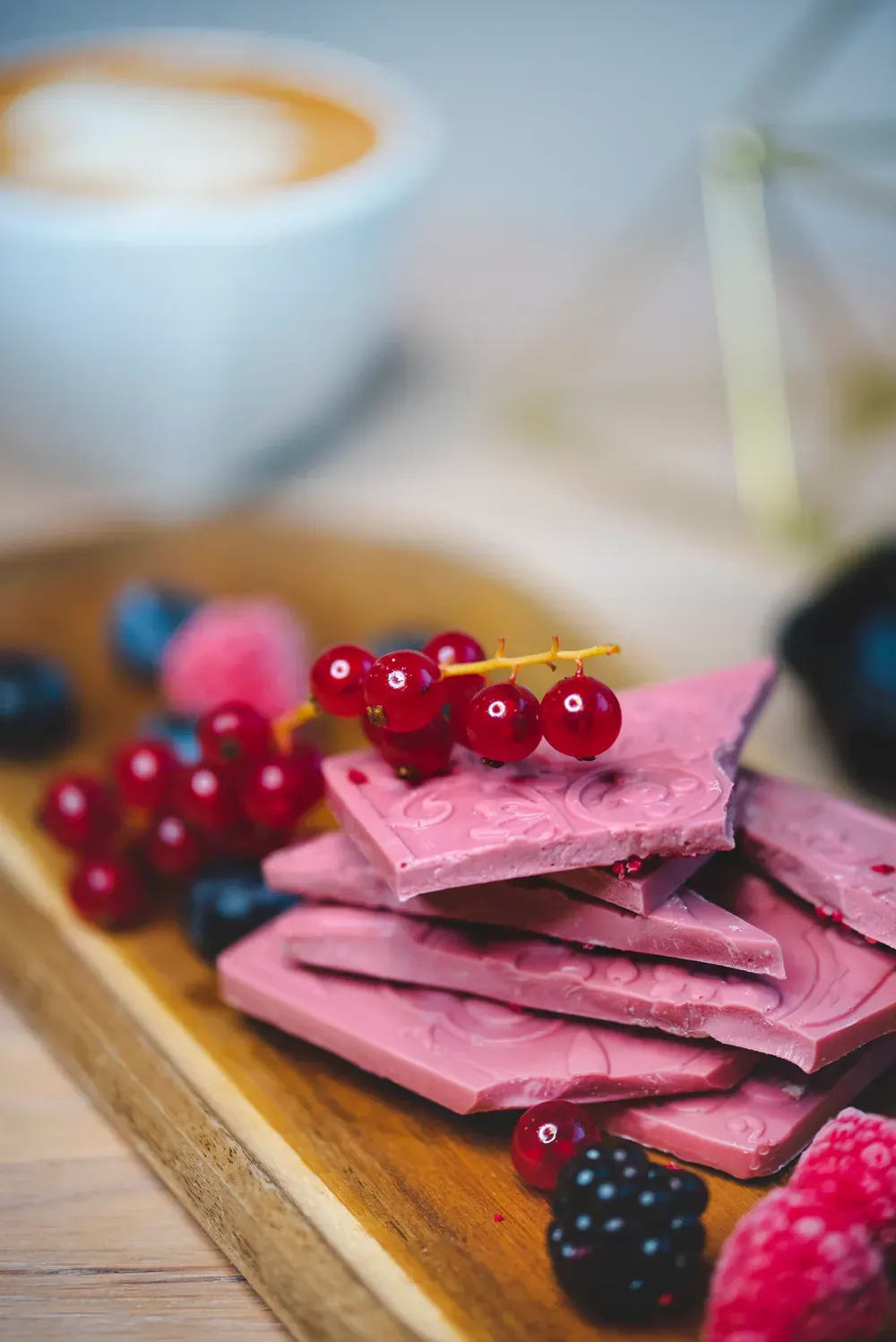 Tablete de ciocolată ruby 47,3% Choco Toni cu coacăze roșii, pe un platou de lemn lângă o cafea